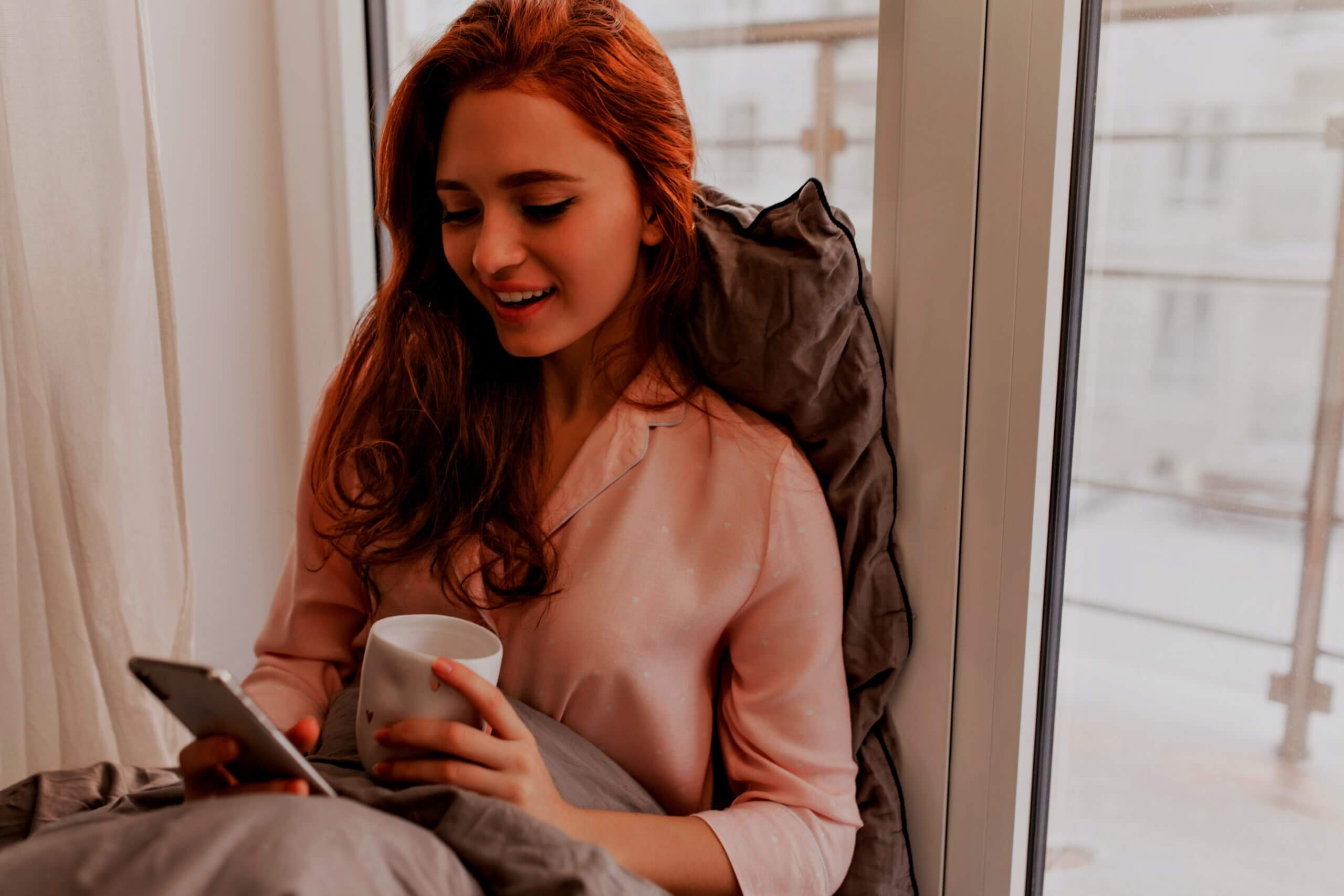 long-haired-ginger-woman-reading-phone-message-in-morning-cute-caucasian-girl-sitting-in-bed-with-cup-of-coffee-and-smartphone-4